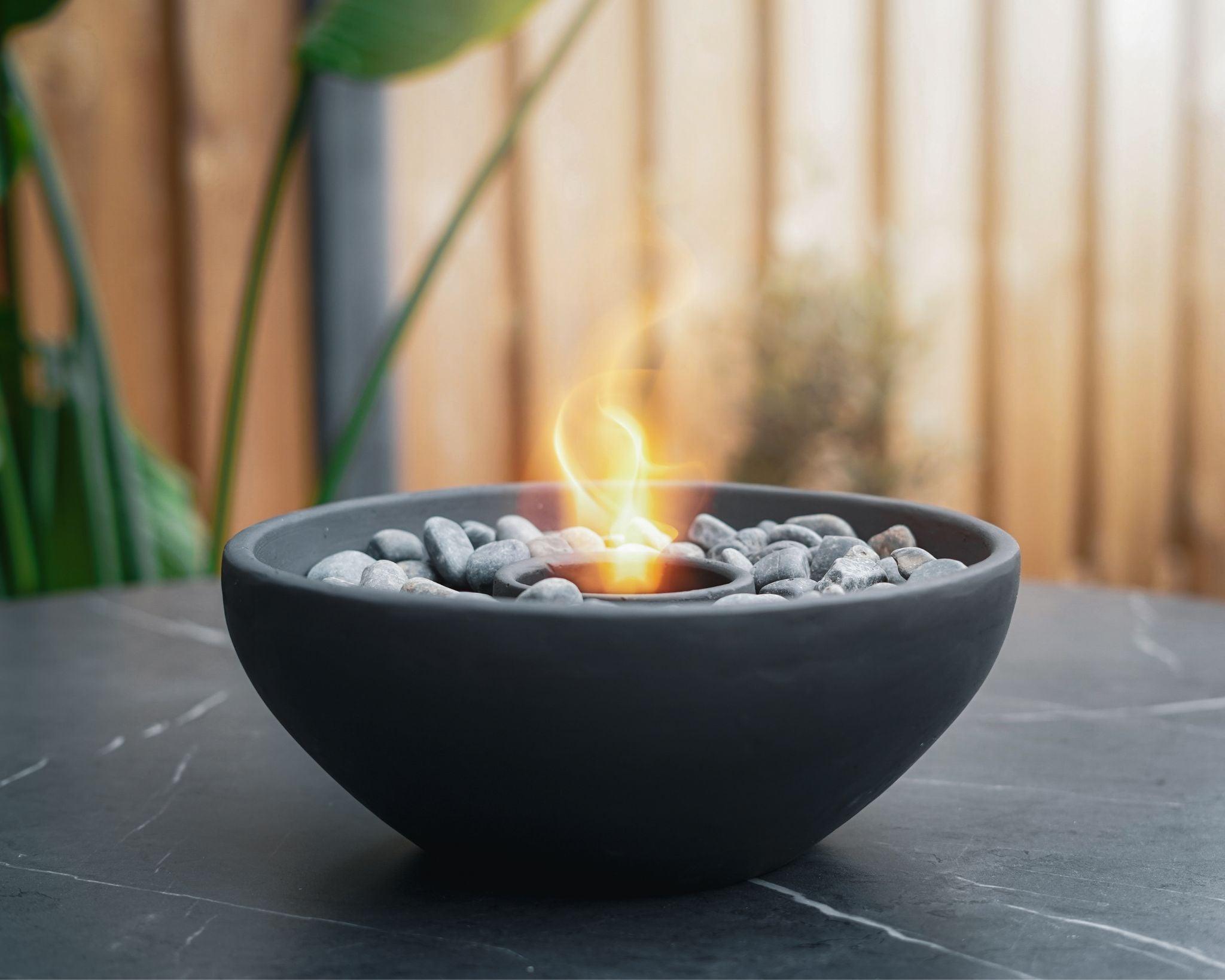 Modern black tabletop fireplace with decorative stones and a dancing flame, placed on a dark outdoor table with a wooden fence and green plants in the background.