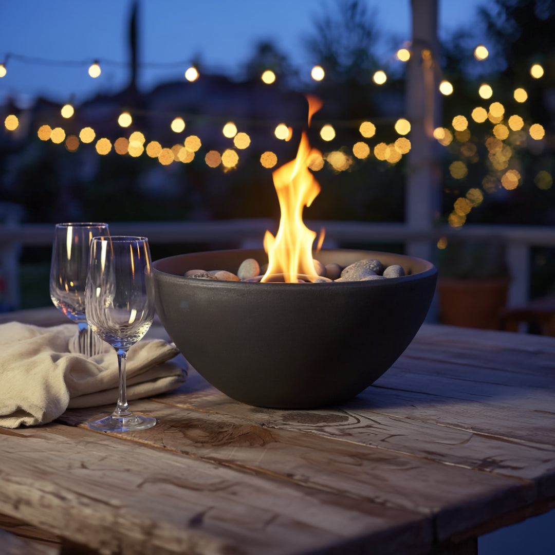 Black round bio ethanol tabletop fire pit on a wooden table with two wine glasses and string lights in the background creating a cozy outdoor evening scene.