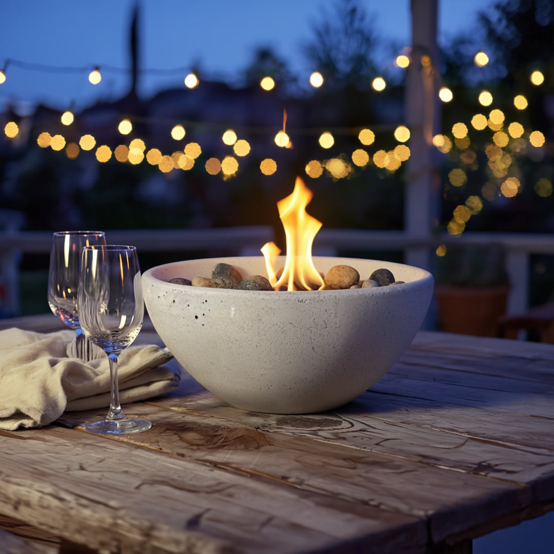 Cream round bio ethanol tabletop fire pit with natural stones on a rustic wooden table, glowing warmly under outdoor string lights during a cozy evening.