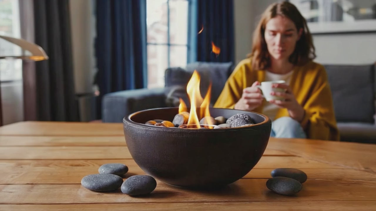 Cosy indoor moment with a bio-ethanol tabletop fire pit burning in focus, while a woman in a mustard yellow cardigan enjoys a warm drink in a softly lit living room with blue curtains.