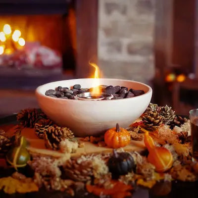 Cream-coloured round bio ethanol tabletop fireplace with flame, surrounded by pinecones and pumpkins in a cosy autumn setting