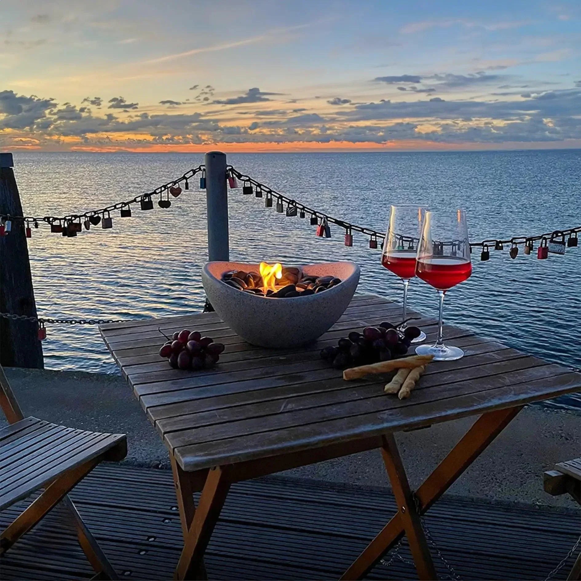 Tabletop bio ethanol fireplace on a wooden outdoor table at sunset, with wine glasses, grapes, and sea view in the background.