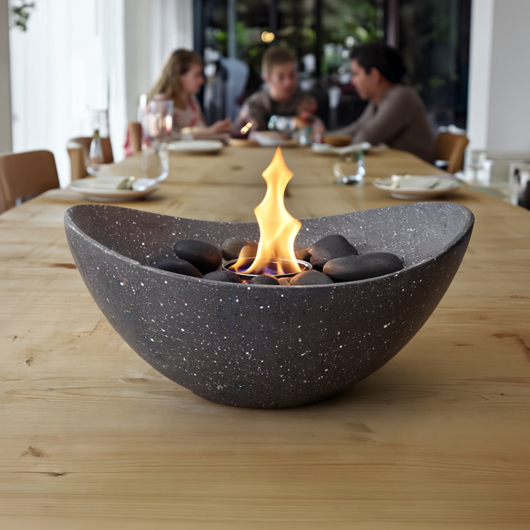 Anthracite oval bio ethanol tabletop fire pit with black stones on a wooden dining table, creating a cozy indoor atmosphere with people in the background