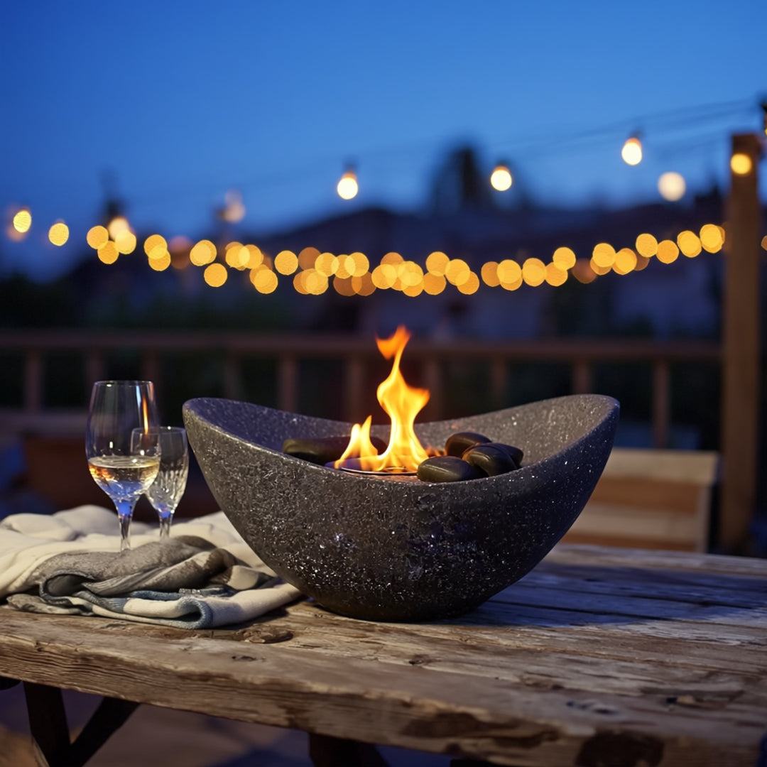 Anthracite oval bio ethanol tabletop fire pit with smooth black stones on a rustic wooden table, glowing warmly under outdoor string lights during a cozy evening.