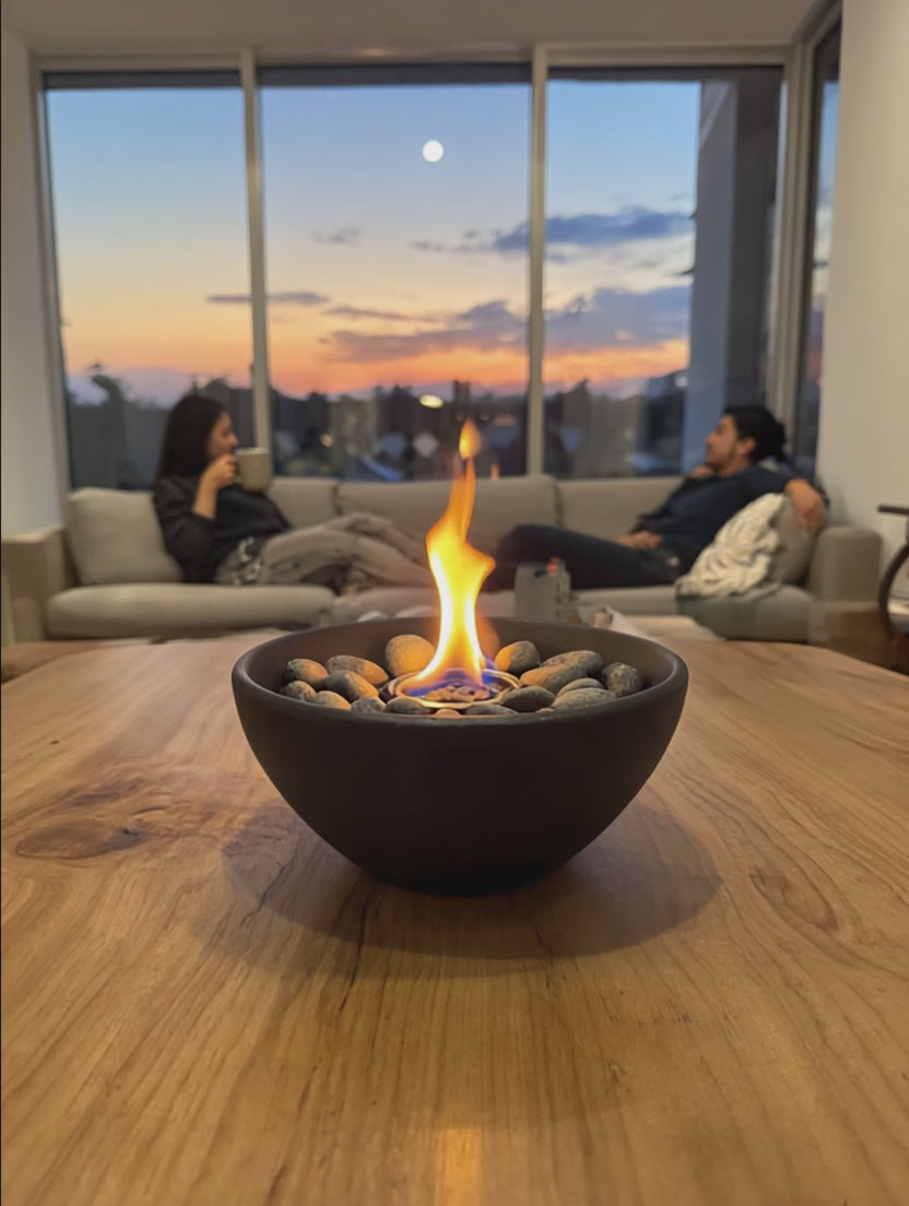 Bio ethanol tabletop fire pit with a calm flame on a wooden table, with two blurred people sitting on a sofa in a warm evening setting.