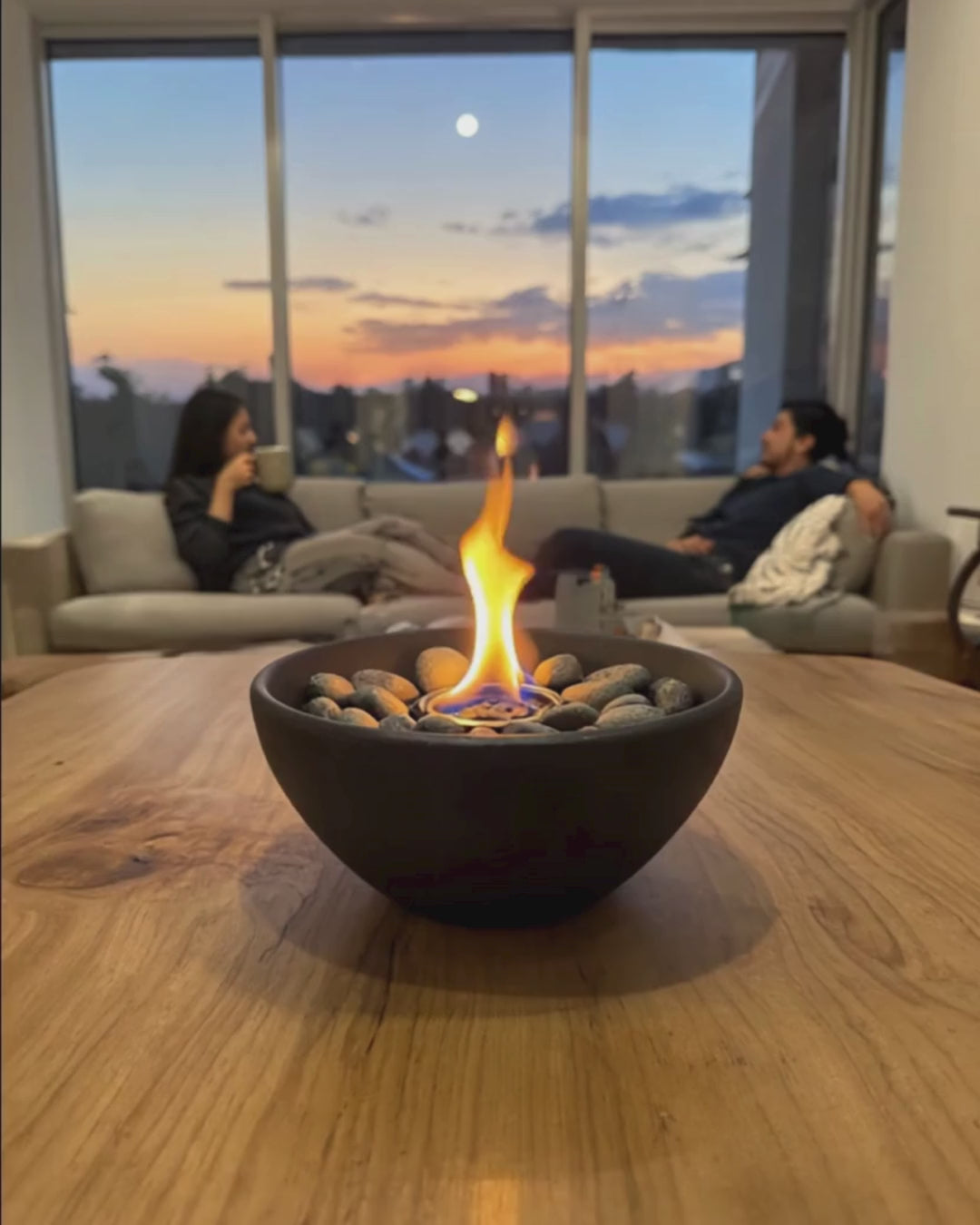 Bio ethanol tabletop fire pit burning on a wooden table with warm indoor evening atmosphere and two people in the background